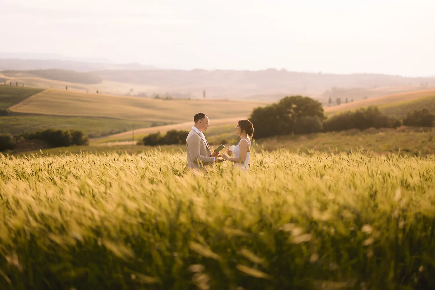 Wide view of Jessica and Chris facing each other for their vow renewal amid tall grain and rolling hills near Pienza.