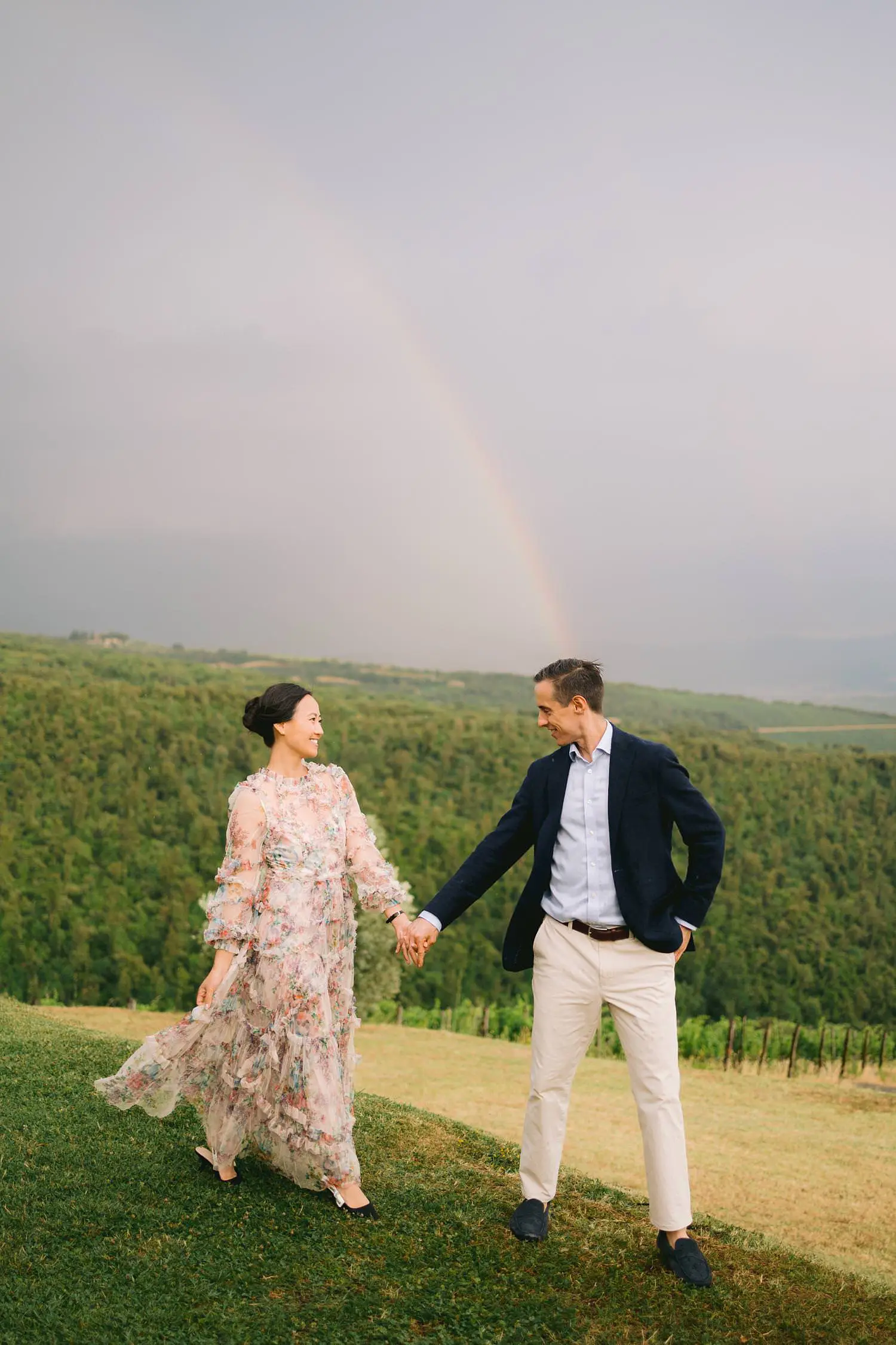 A newly engaged couple holds hands beneath a rainbow arching above the vineyard at Castiglion del Bosco.
