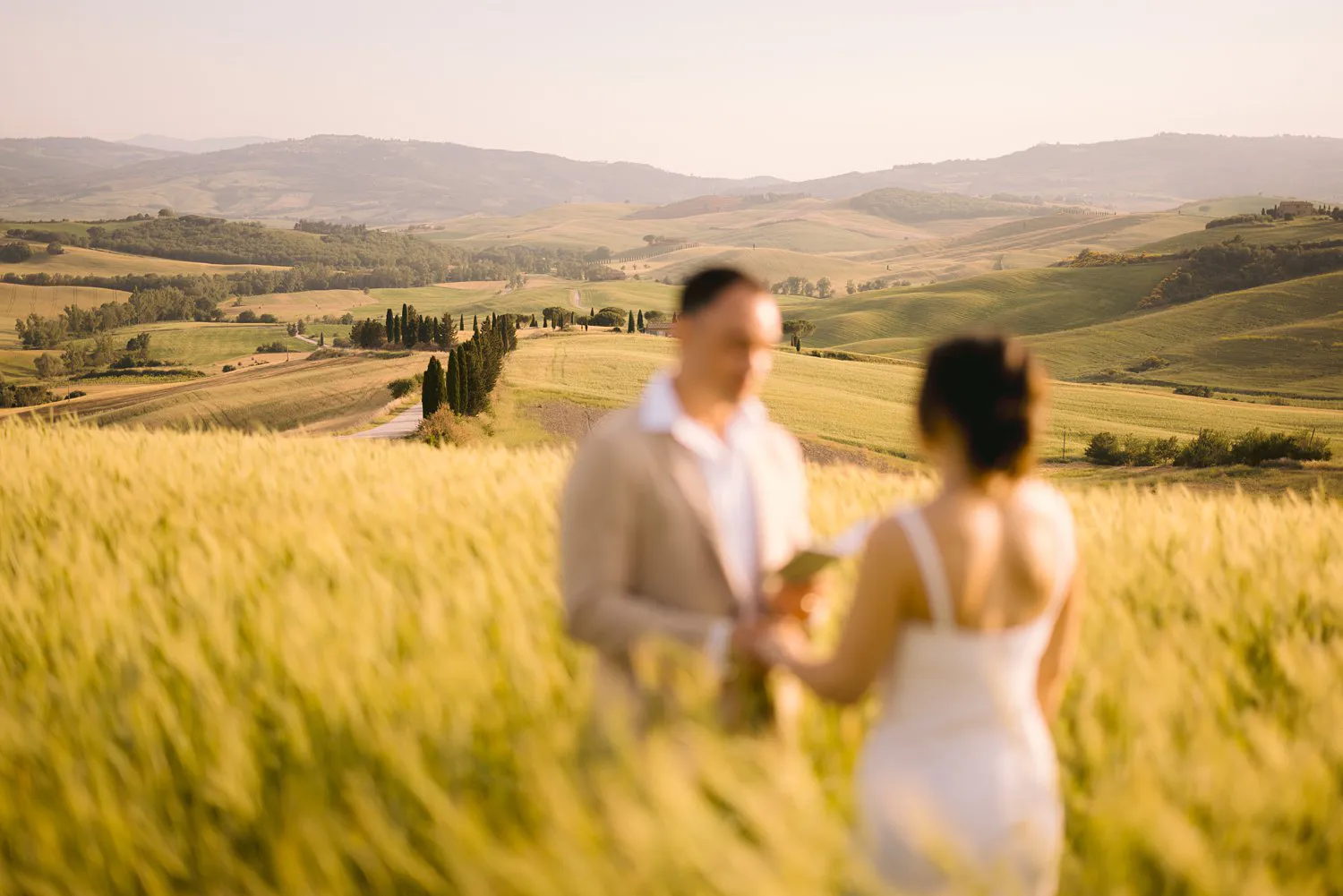 Soft-focus view of Jessica and Chris holding their written vows, with a winding cypress-lined road in the valley behind them.