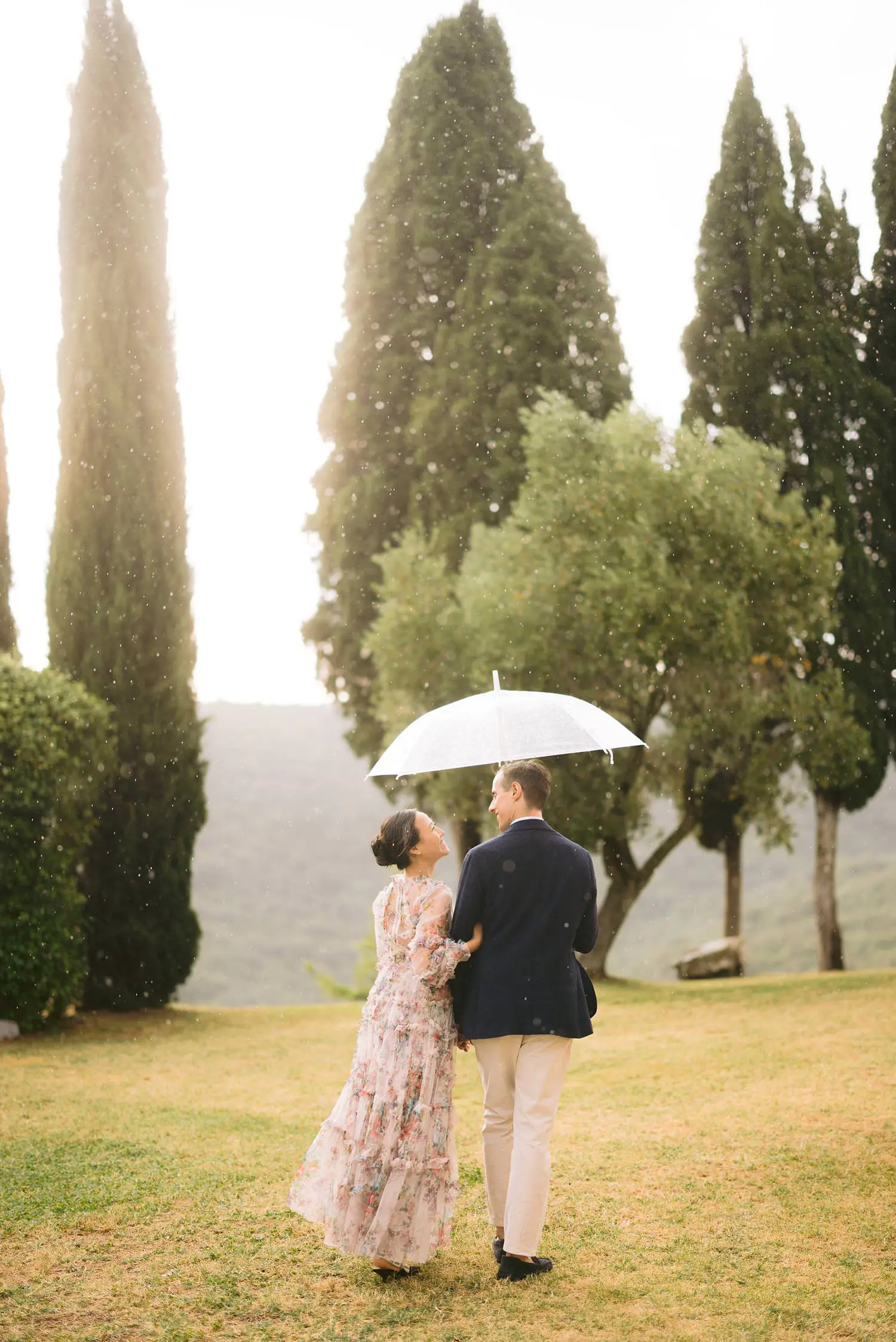 A newly engaged couple stands beneath a transparent umbrella, framed by tall cypresses at Capanna House in Val d’Orcia.