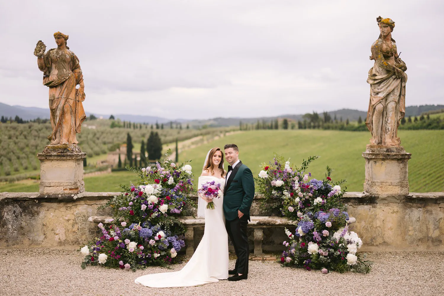 Kathryn holds her bouquet beside Kevin between floral arrangements and stone statues in Villa Corsini's garden, with the Tuscan hills behind them.