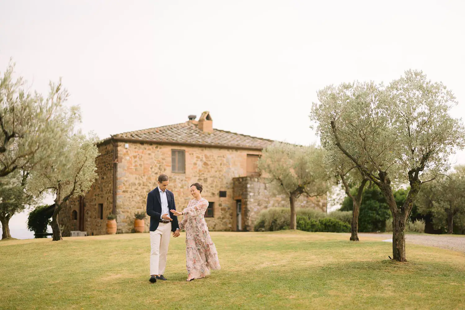 A couple walks across the lawn in front of Capanna House, its stone facade framed by olive trees.