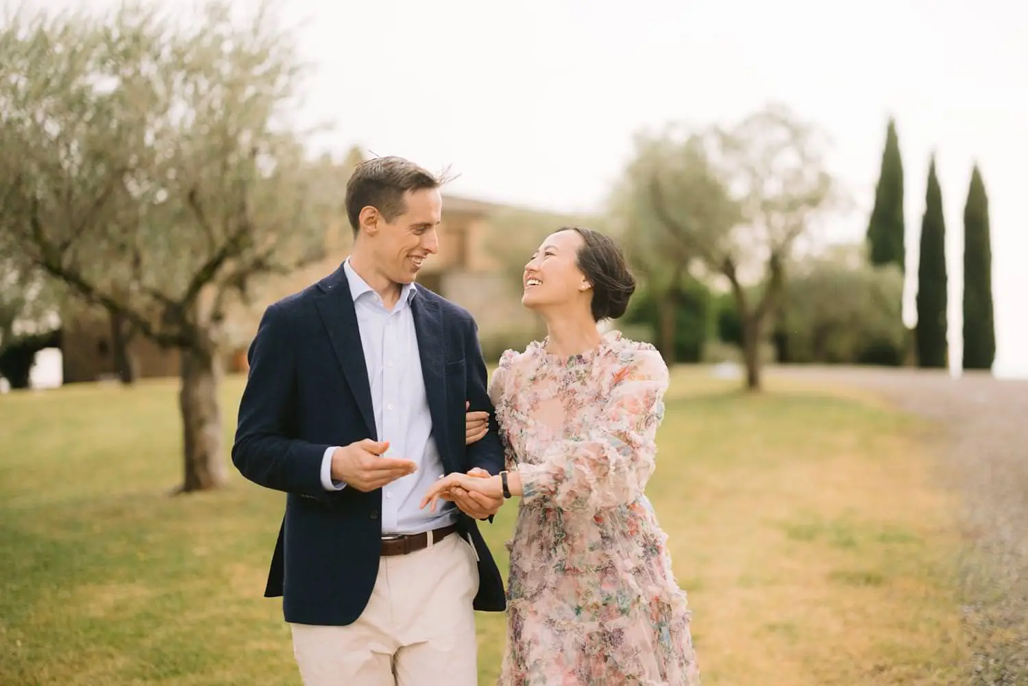 A woman smiles up at her partner as they walk among olive trees and cypresses on the grounds of Capanna House.