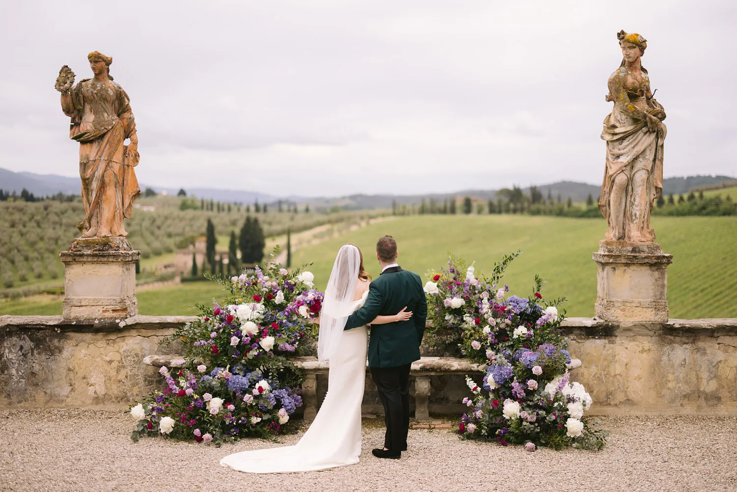 Rear view of Kathryn and Kevin standing arm in arm between floral arrangements and stone statues, looking across the countryside at Villa Corsini.