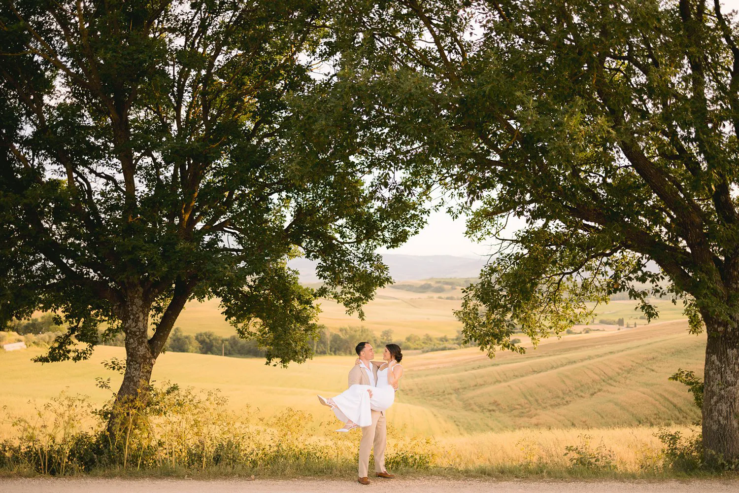 Chris carries Jessica beneath two leafy trees beside a country road, with the fields of Val d'Orcia stretching behind them.