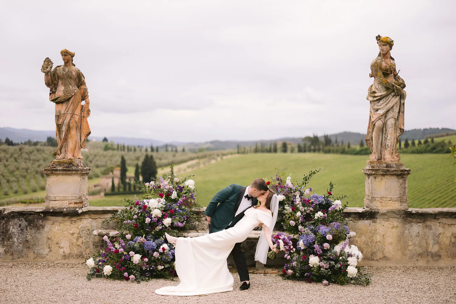 Kevin dips Kathryn backwards into a kiss between purple floral arrangements and stone statues in the garden at Villa Corsini.