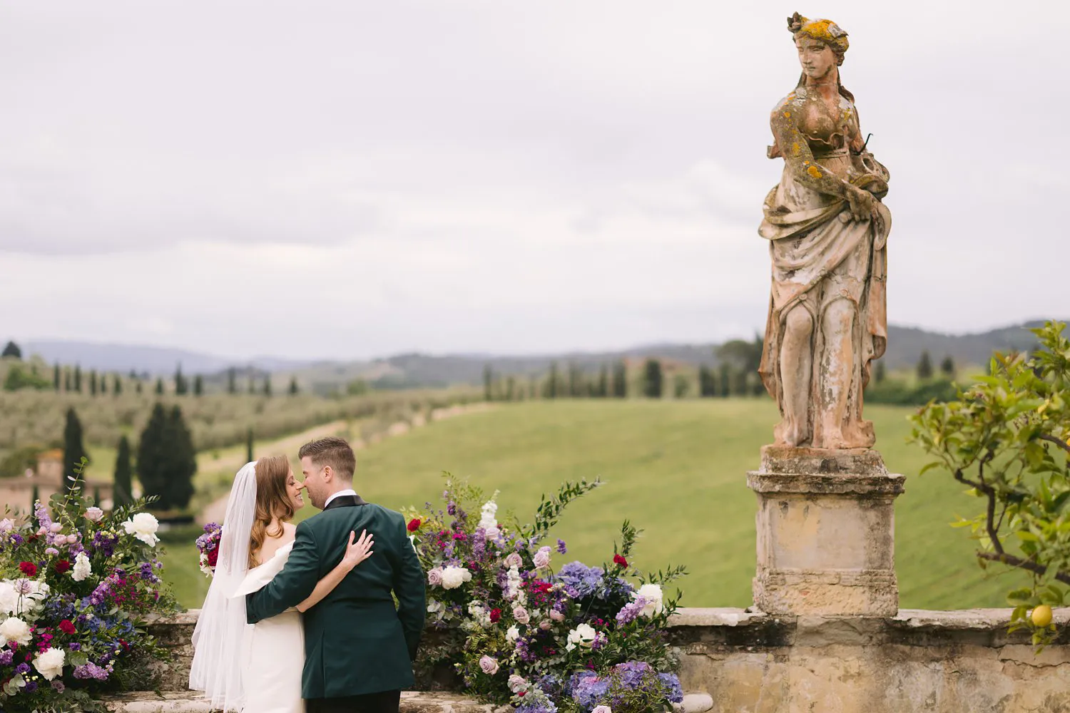 Kathryn and Kevin kiss among purple and pink flowers beside a weathered stone statue overlooking the Tuscan countryside at Villa Corsini.