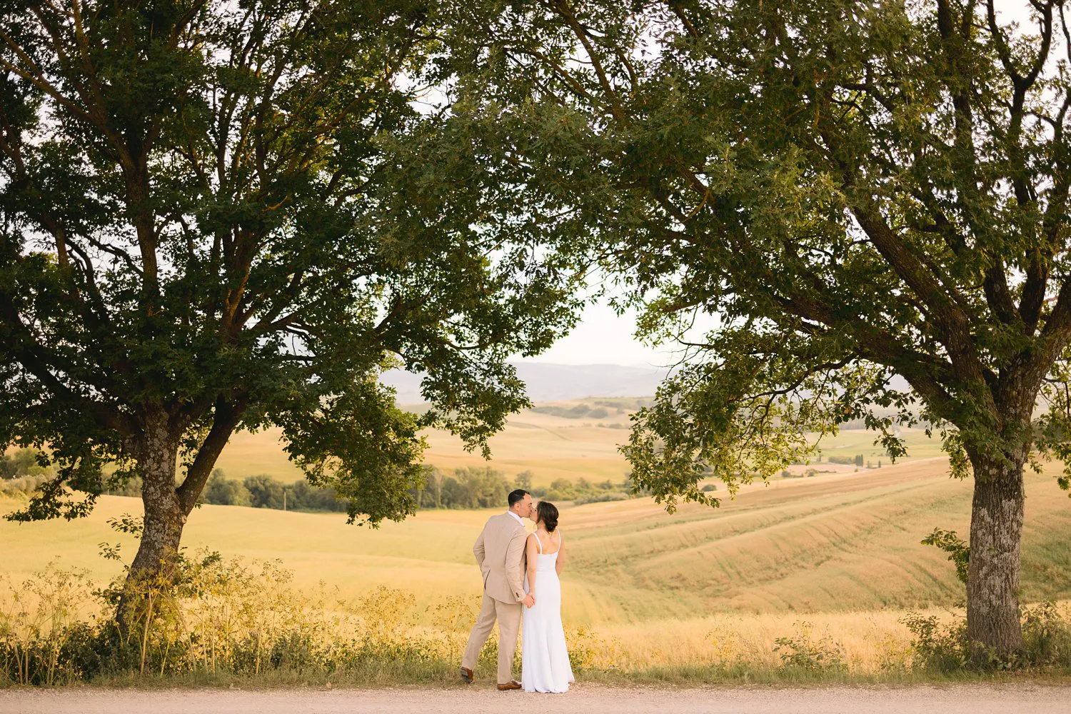 Jessica and Chris stand close beneath two leafy trees beside a country road, framed by sunlit fields near Pienza.