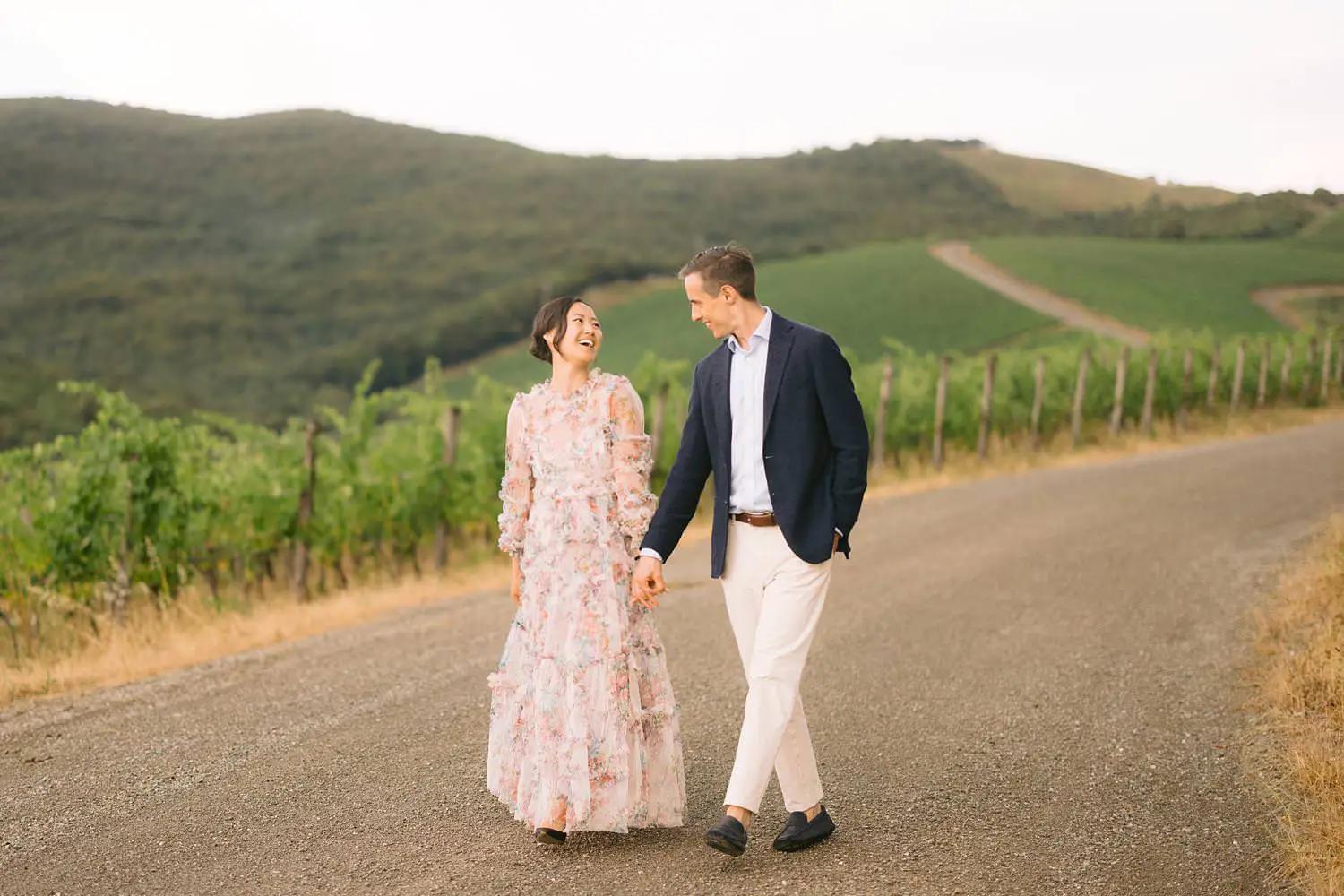 A newly engaged couple walks hand in hand along a quiet road beside green vineyards and rolling Tuscan hills.