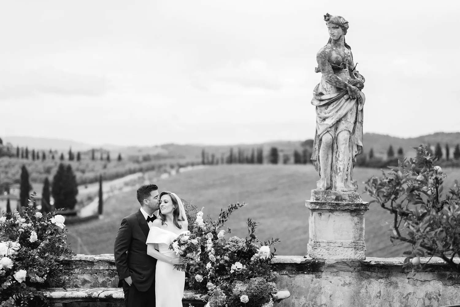 Black-and-white portrait of Kathryn leaning against Kevin beside a floral arrangement and a tall stone statue in the gardens of Villa Corsini.