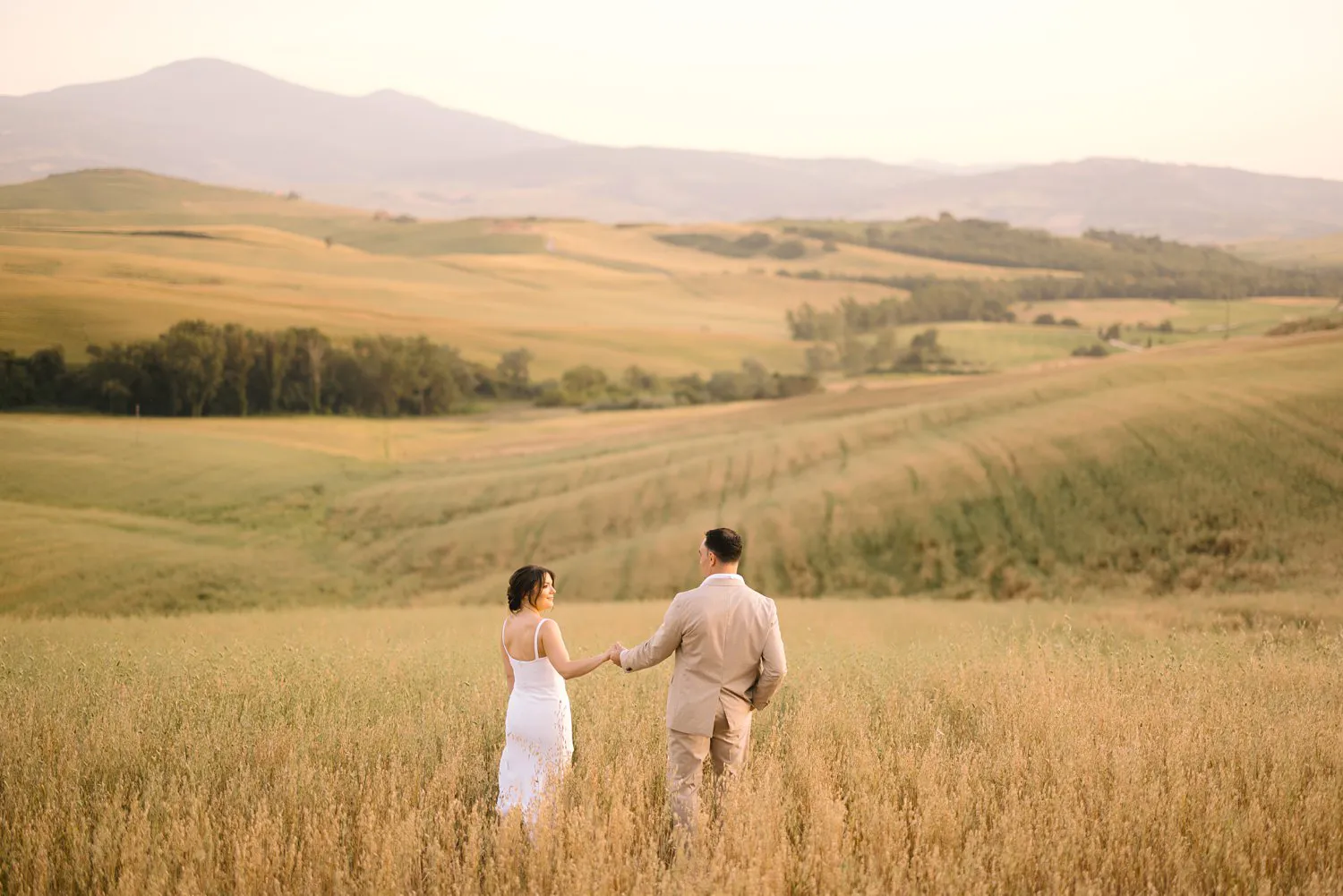 Seen from behind, Jessica and Chris walk hand in hand through tall grain toward the layered hills of Val d'Orcia.