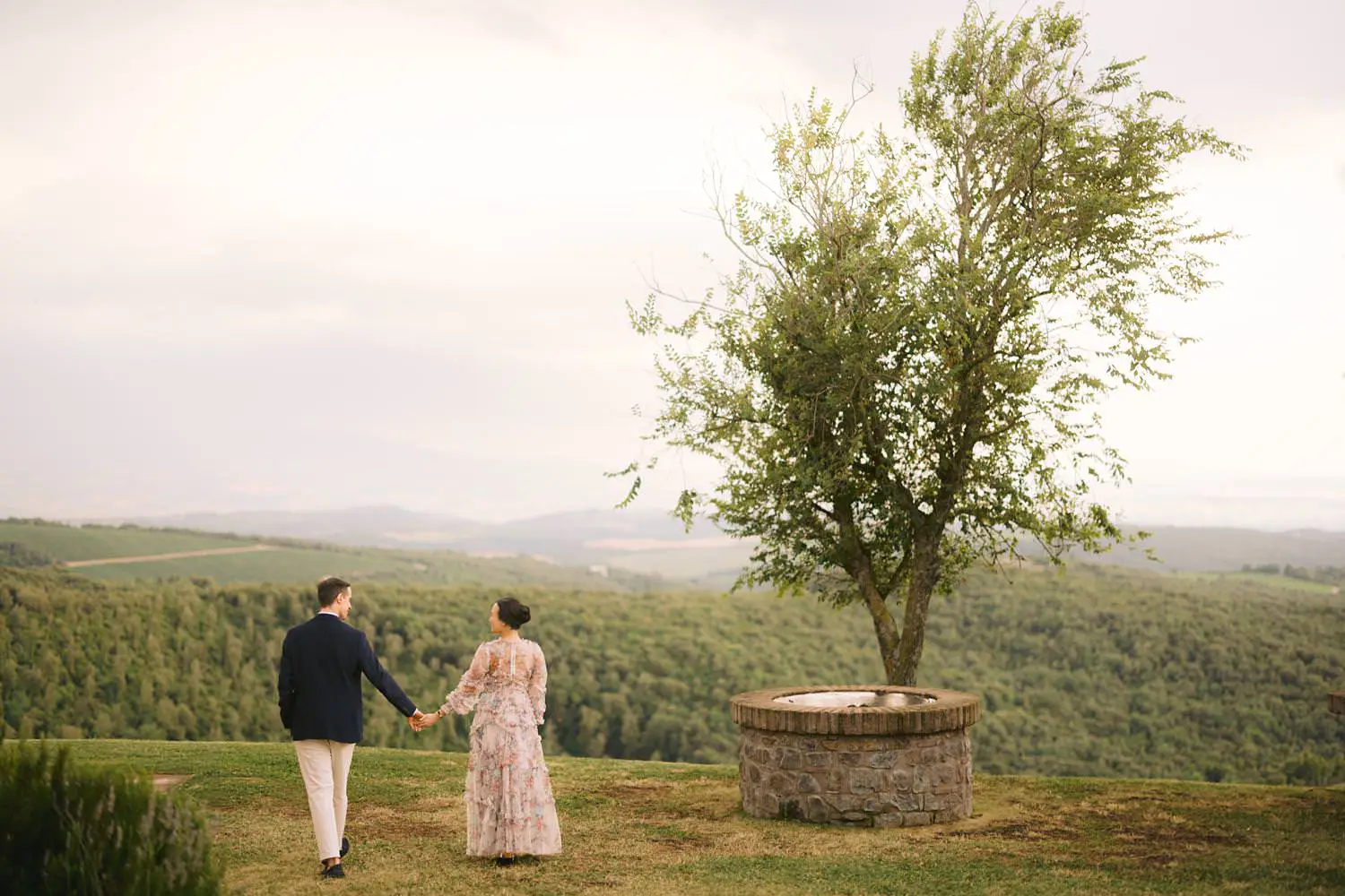 Hand in hand, couple walk toward the vineyard views at Capanna House, beside a tree and a circular stone well.