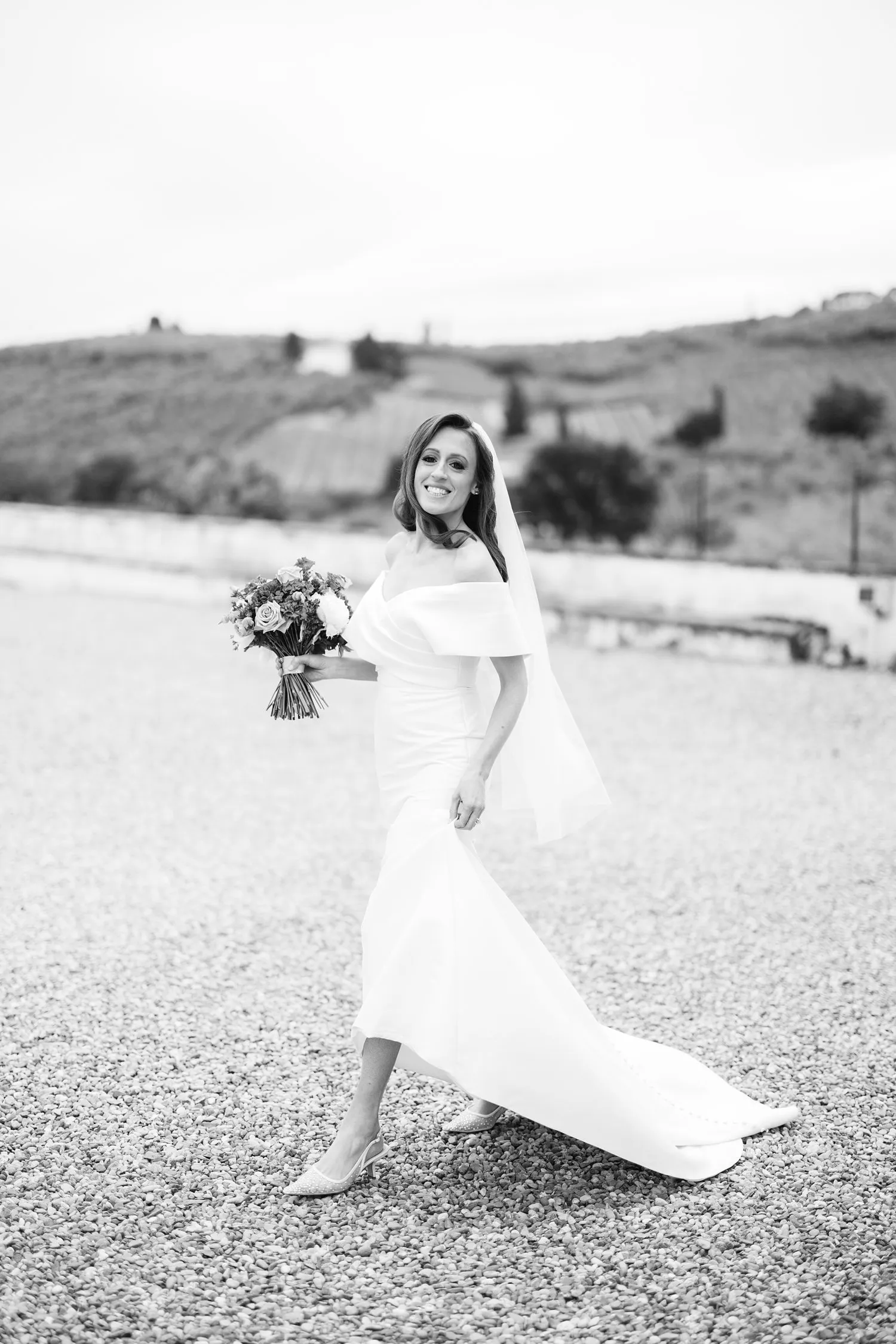Black-and-white candid portrait of Kathryn smiling as she walks across the gravel garden at Villa Corsini, carrying her bouquet with her gown in motion.