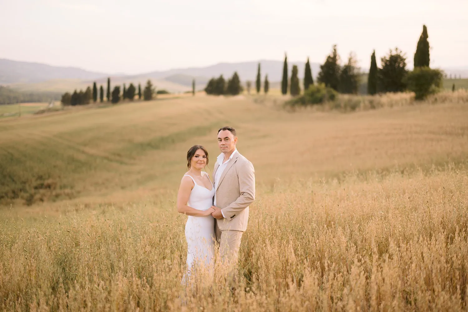 Jessica and Chris stand together in a field near Pienza, her white dress and his beige suit framed by grain and distant cypresses.