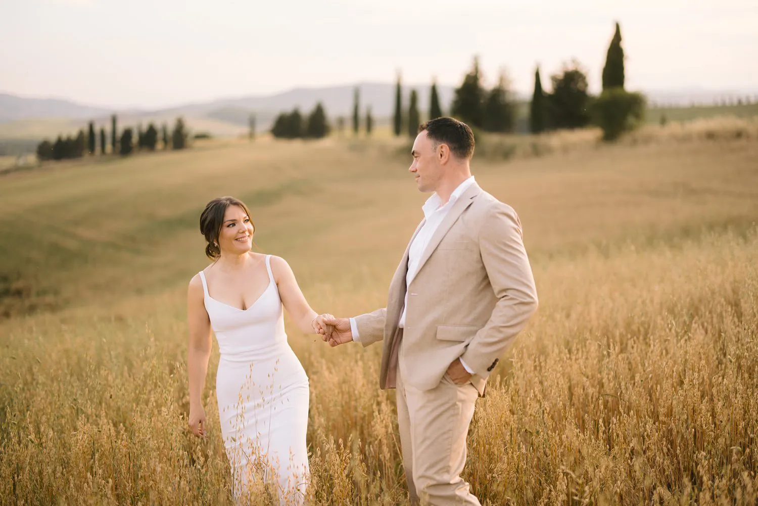 Jessica and Chris look at each other while holding hands in a field of grain, with cypress trees and hills behind them.