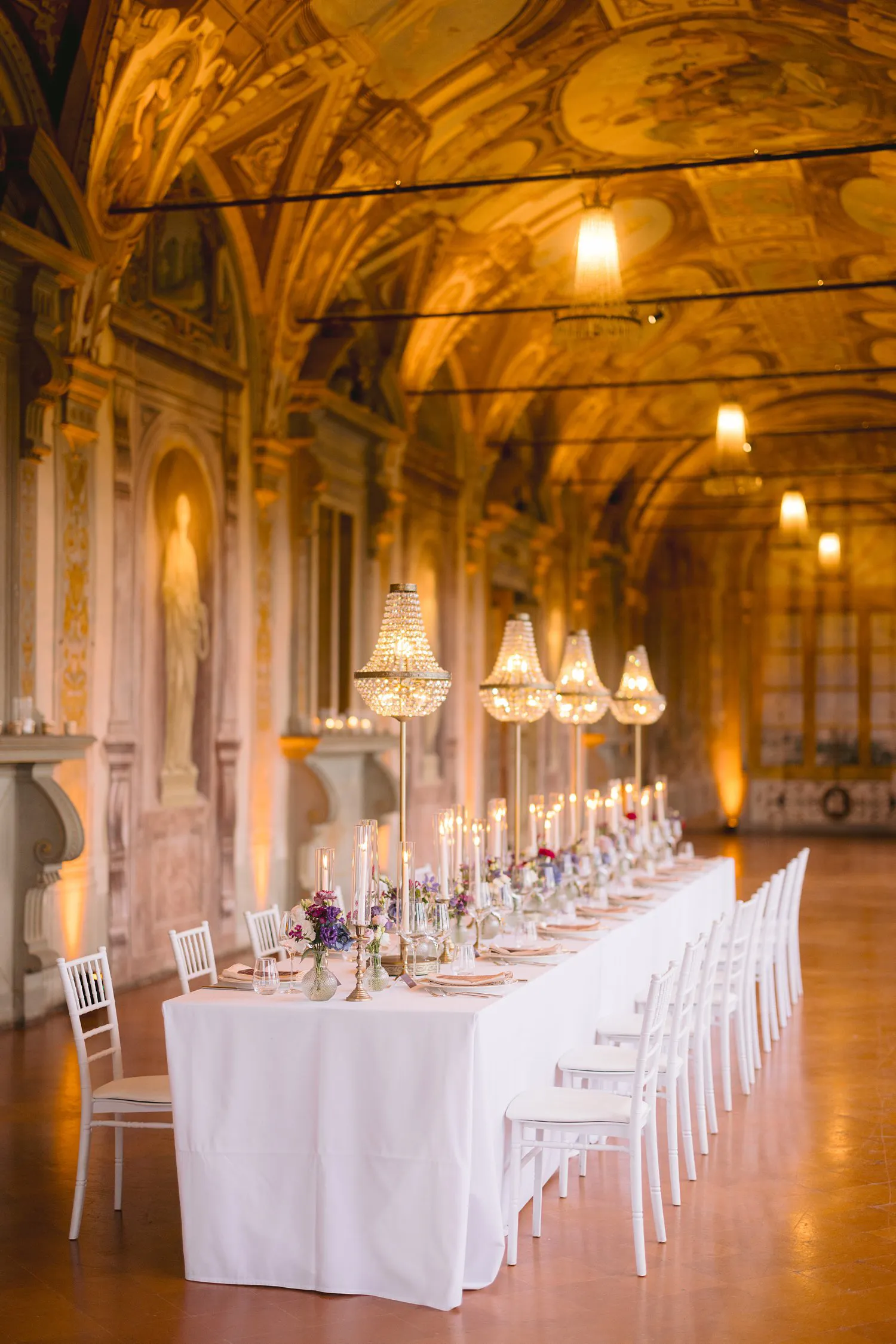 A long white wedding table with candles, flowers and suspended chandeliers stands beneath the frescoed ceiling of the Gallery at Villa Corsini.