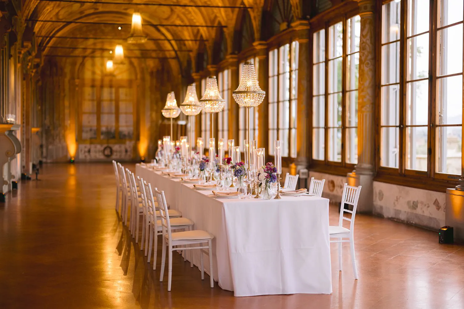 Wide view of the frescoed Gallery at Villa Corsini, with one long wedding table, white chairs, candles and chandeliers beside tall windows.