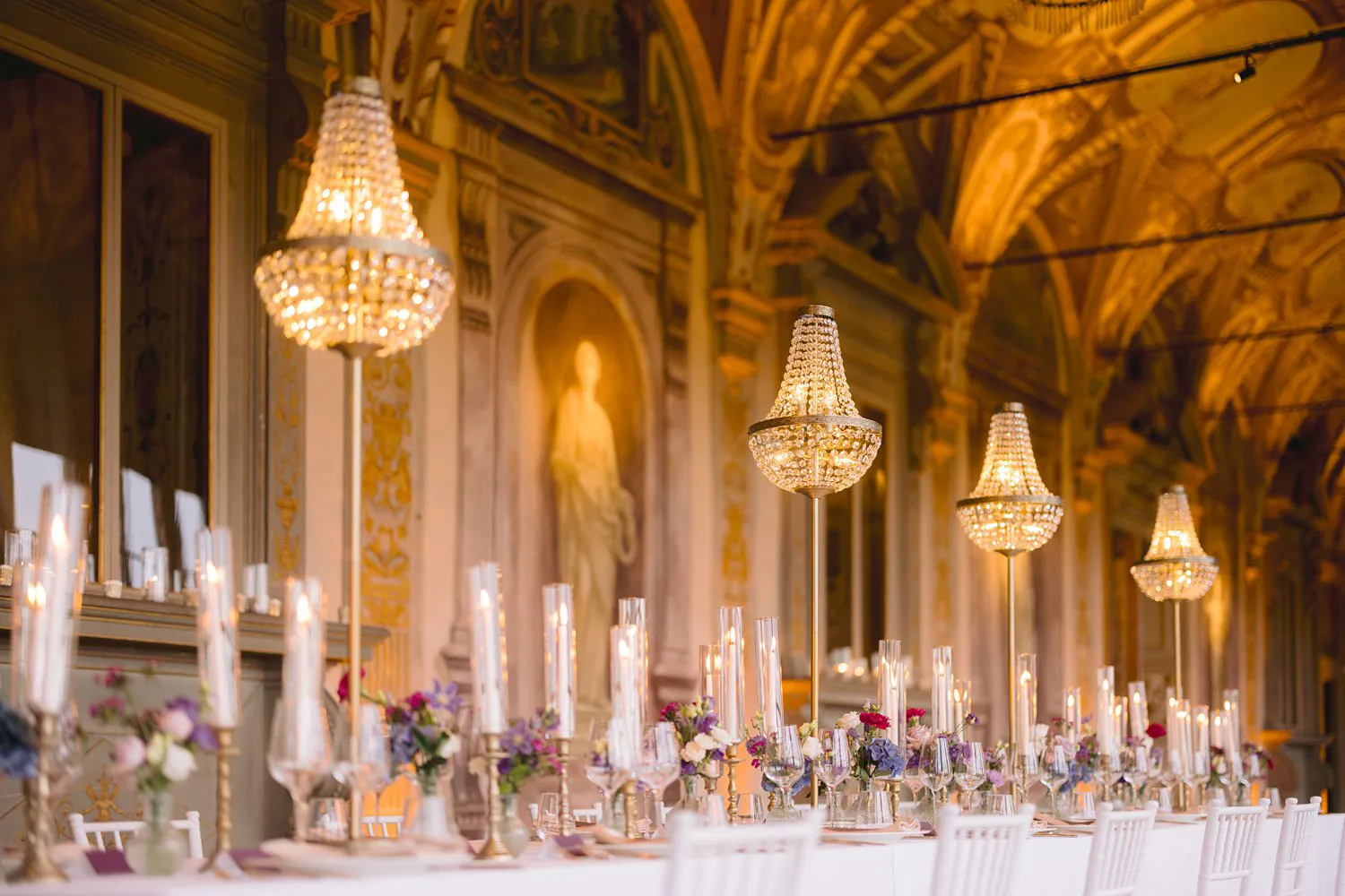 Suspended crystal chandeliers hang over a wedding table with candles and flowers beneath the frescoed ceiling of the Gallery at Villa Corsini.