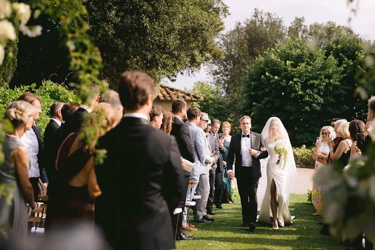 Bride and her father walk down the garden aisle at Villa Le Fontanelle as guests look on.