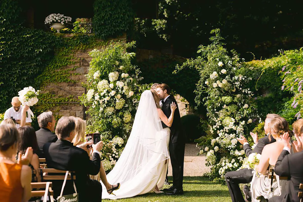 Madison and Travis share their first kiss beneath white floral arches as guests applaud at Villa Le Fontanelle in Florence.