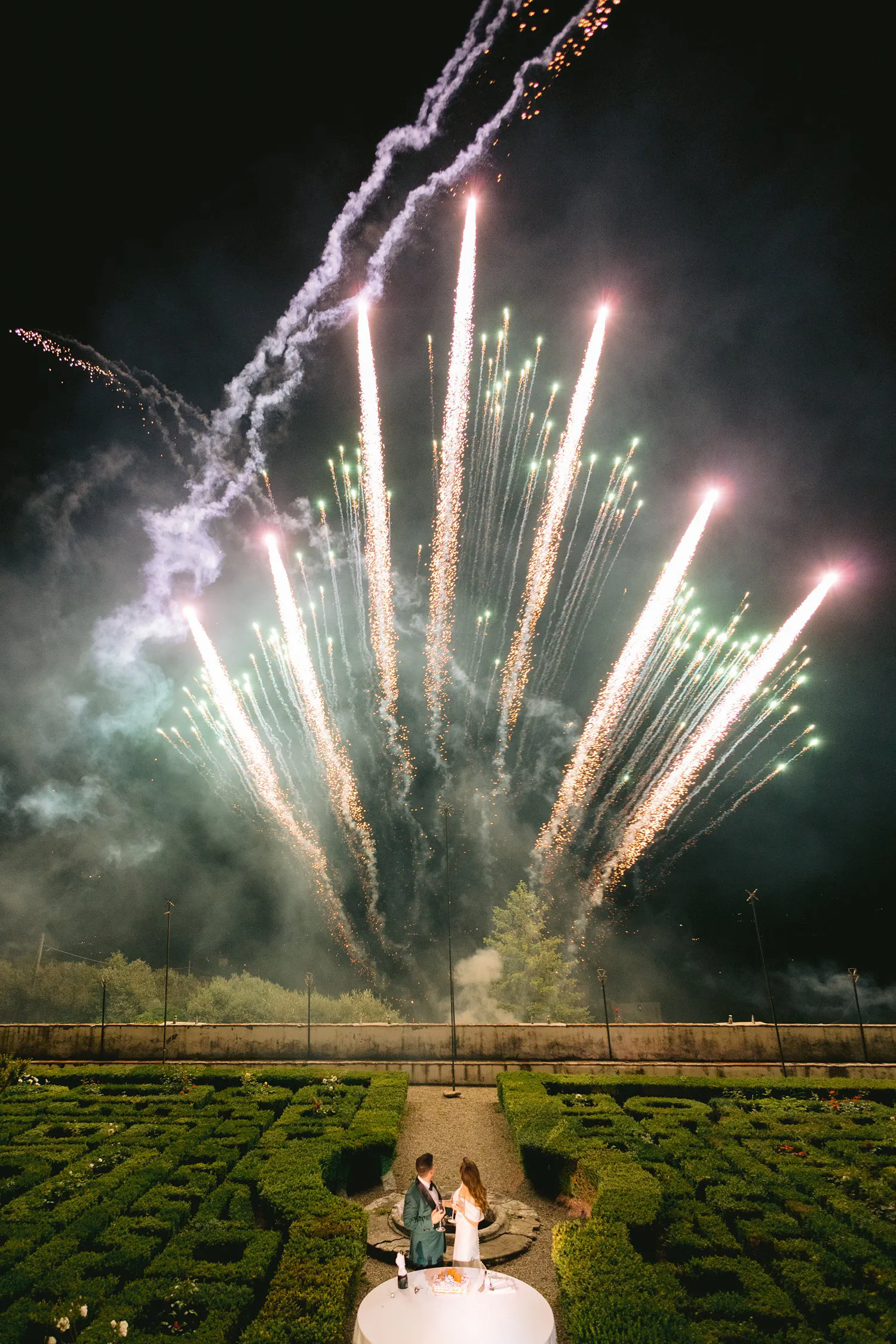 Fireworks rise above the geometric hedges of Villa Corsini's garden at night, with Kathryn and Kevin standing together in the foreground.