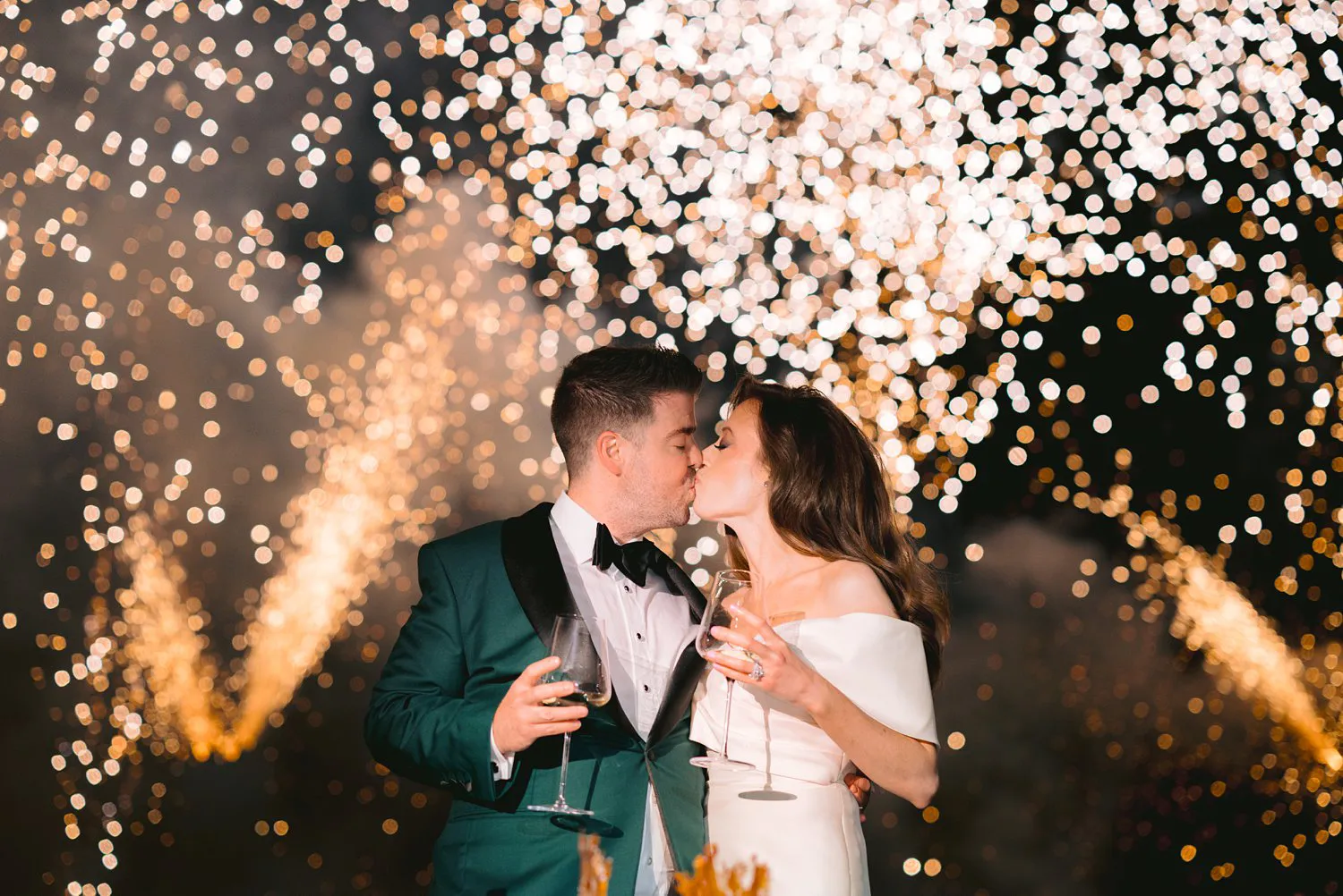 Kathryn and Kevin kiss while holding champagne glasses, with bright golden fireworks filling the background at their Villa Corsini wedding.