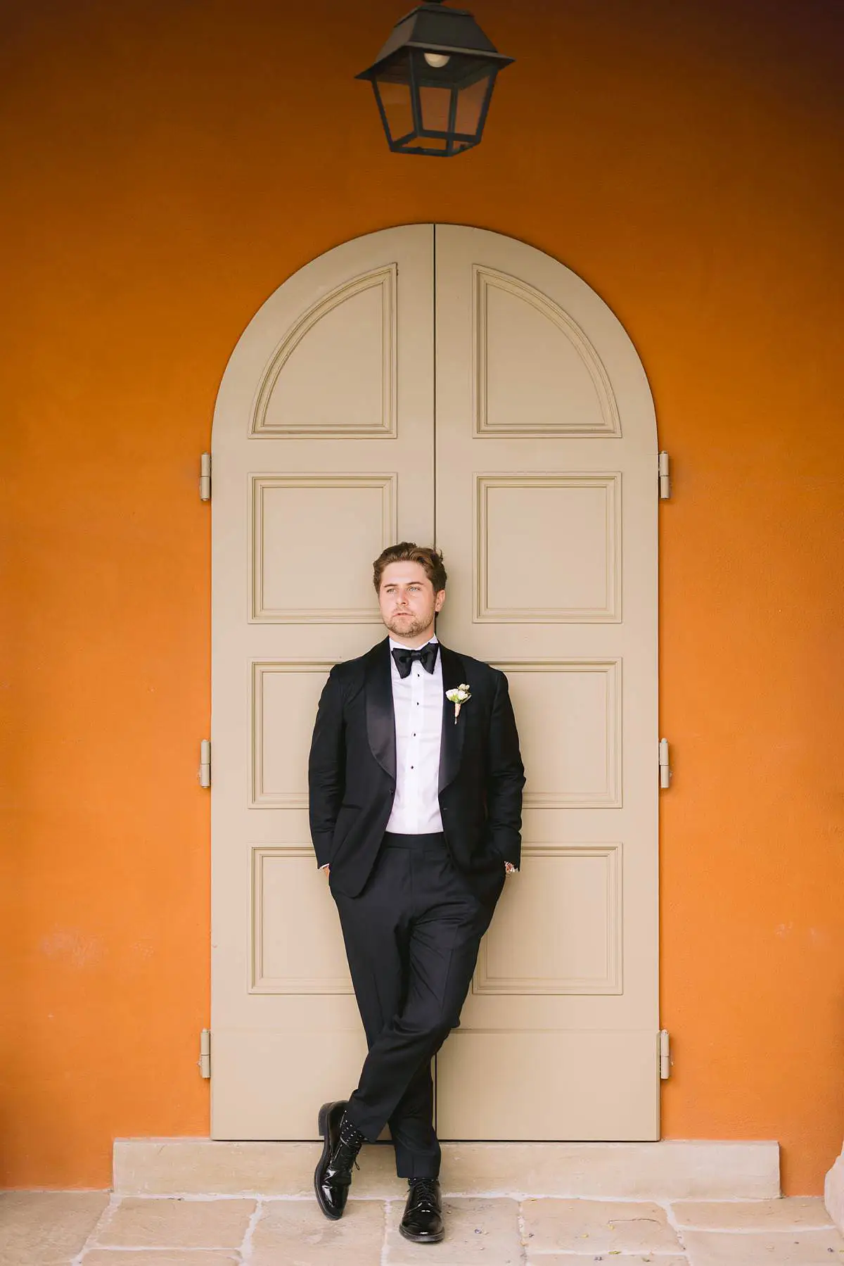 Groom in a tuxedo stands by an orange door at Villa Le Fontanelle.