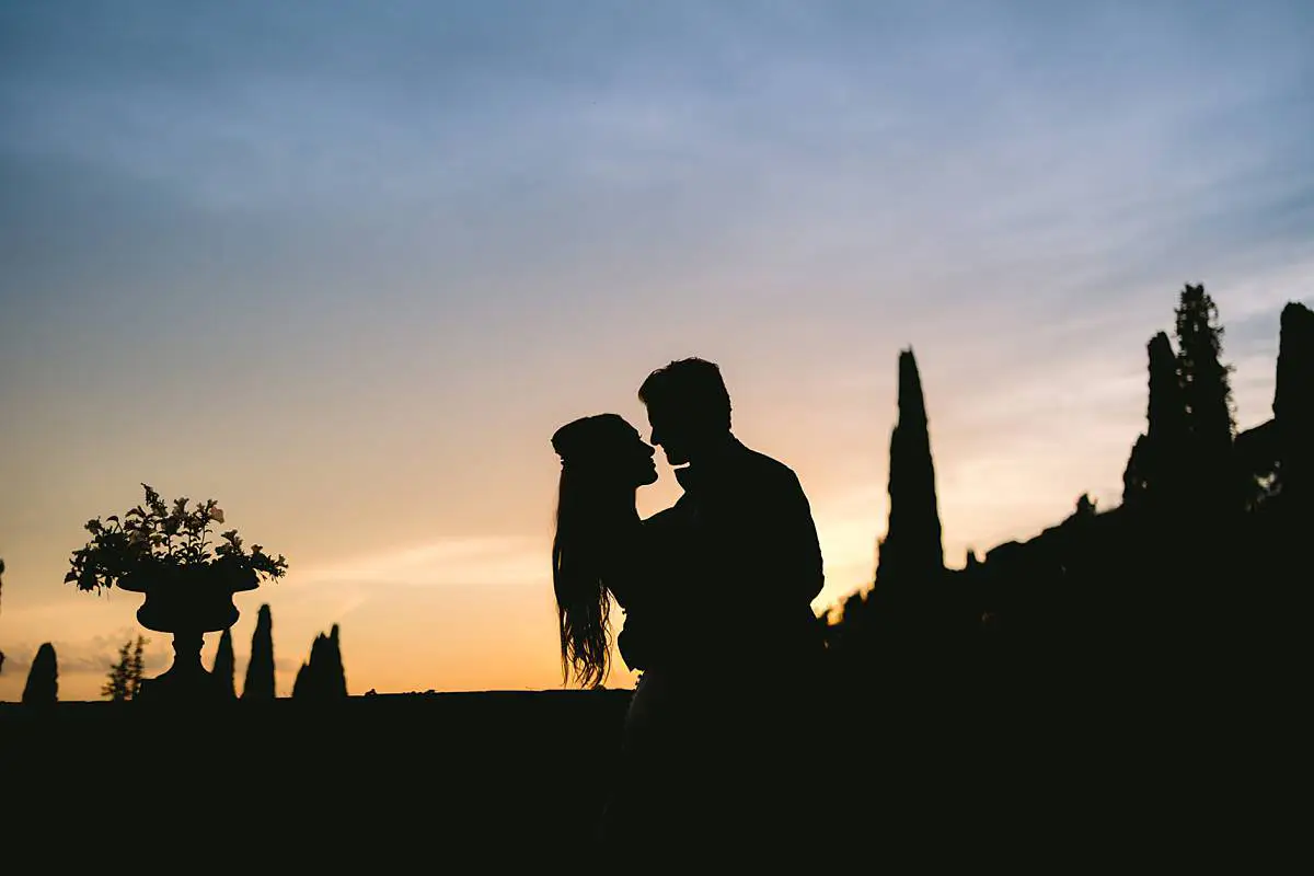 Newlyweds kiss in silhouette at sunset among the cypress trees of Villa Le Fontanelle.